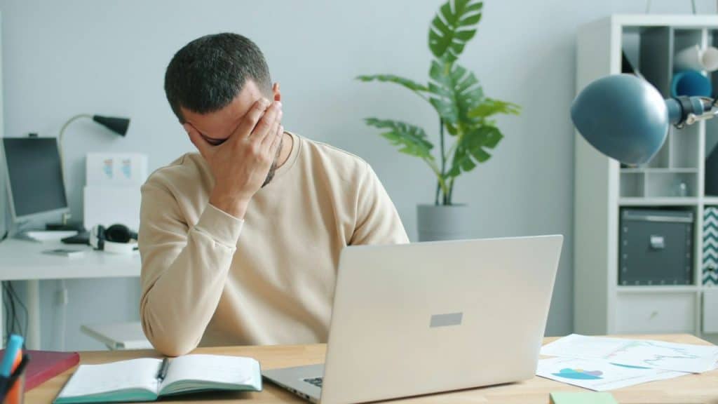 A frustrated man covers his face with his hand while sitting at a desk with a laptop.