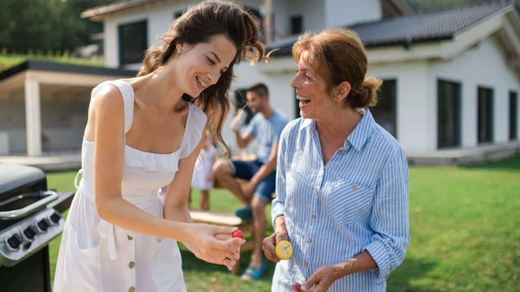A mother-in-law and daughter-in-law are grilling vegetables in the garden while a man is sitting in the background.