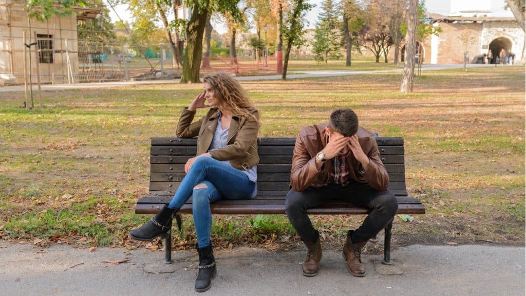 A distressed couple sitting apart on a park bench in autumn, with the woman looking away and the man holding his head in his hands.