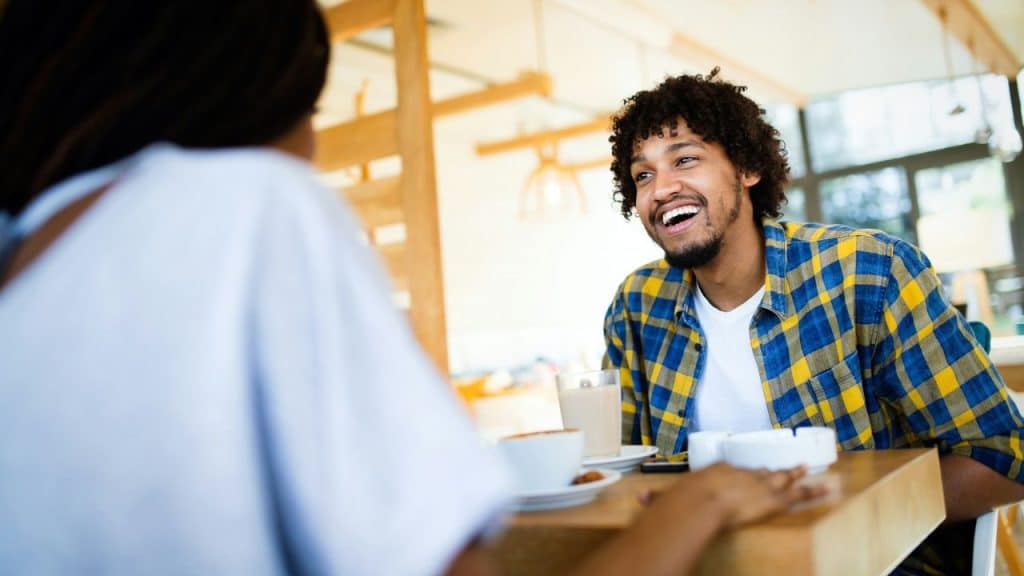 A curly-haired man in a yellow and blue plaid shirt laughs across a table.