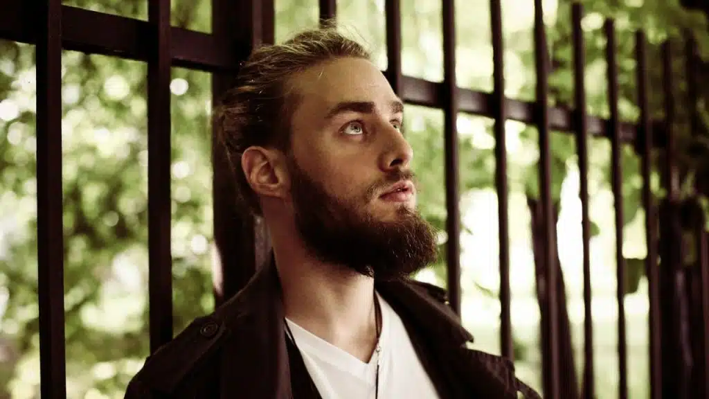 A thoughtful bearded man is looking up while leaning against a dark metal fence.