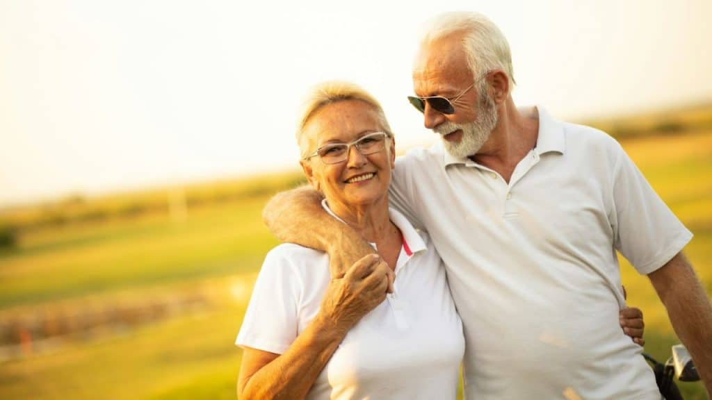 An older couple, both in white shirts, stands outdoors with the man's arm around the smiling woman.