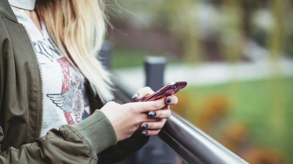 A close-up of a young woman's hands with dark nail polish, holding and looking at a pink smartphone.