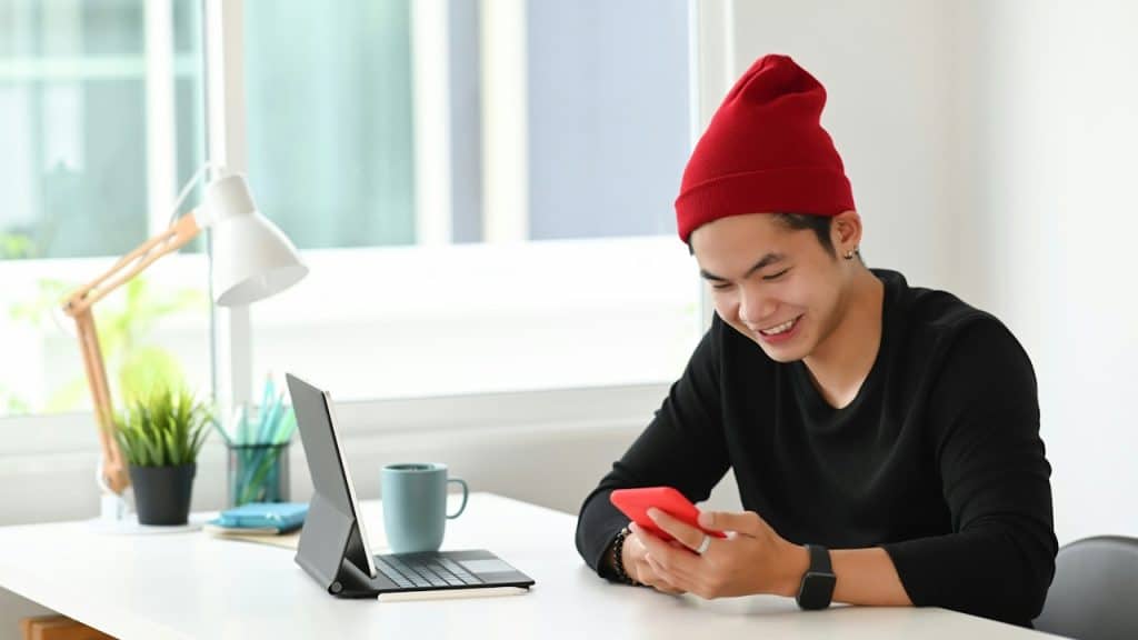 A youngย man in a red beanie smiles while looking at his red smartphone at a desk with a laptop.
