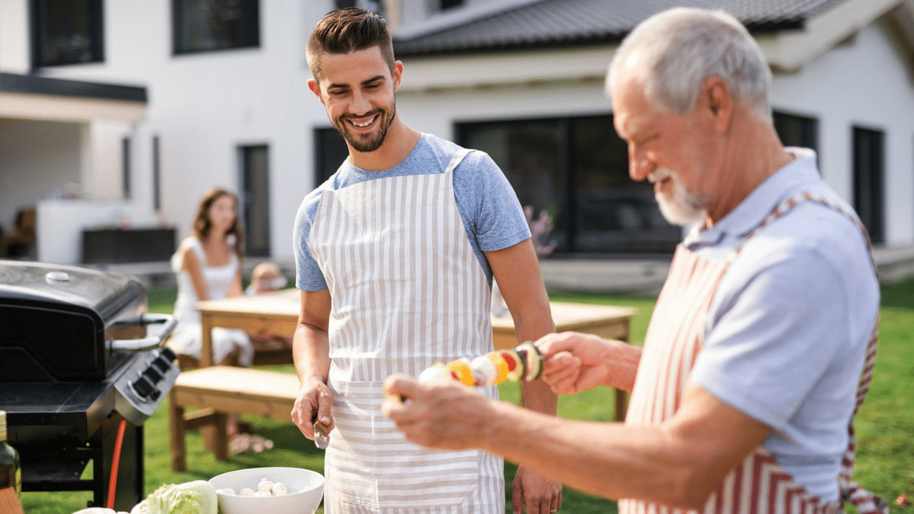 Two men in aprons preparing food outdoors next to a grill; one holds a kebab.