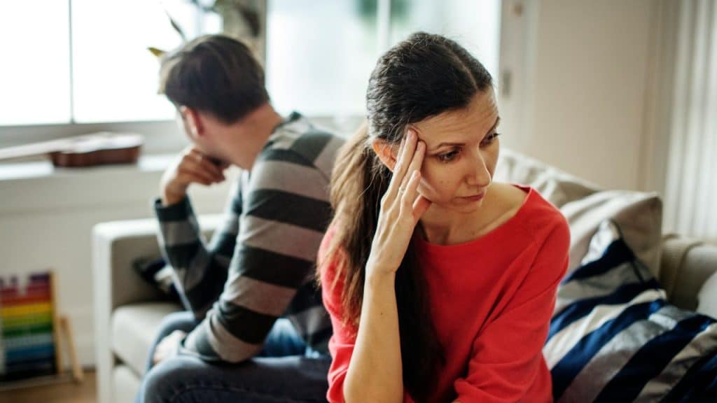 An upset woman in a red shirt sits with her hand on her head, facing away from a man.