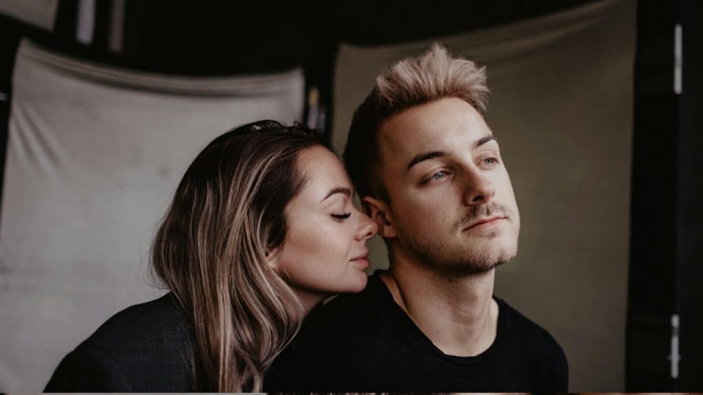A woman with her eyes closed leans her head affectionately on a man's shoulder, who is looking thoughtfully to the side.