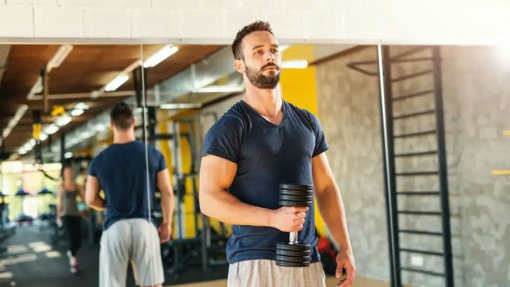 A man with a beard and dark shirt holding a dumbbell in a gym, looking determined.
