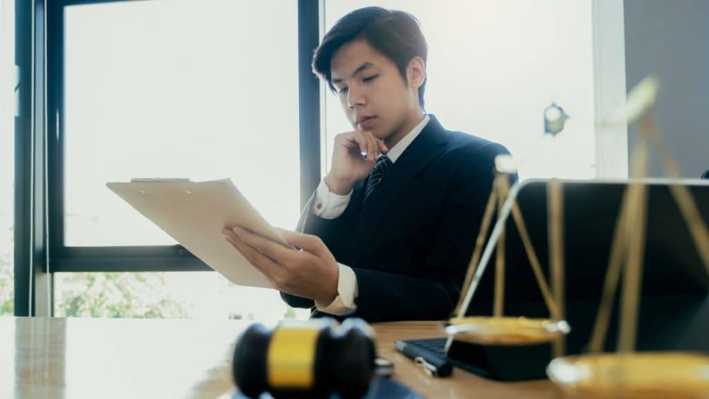 Young man in a suit reviewing a document on a clipboard with a gavel and scales of justice on the table.