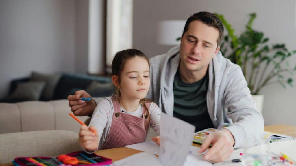 Man assisting a young girl with homework at a table indoors.