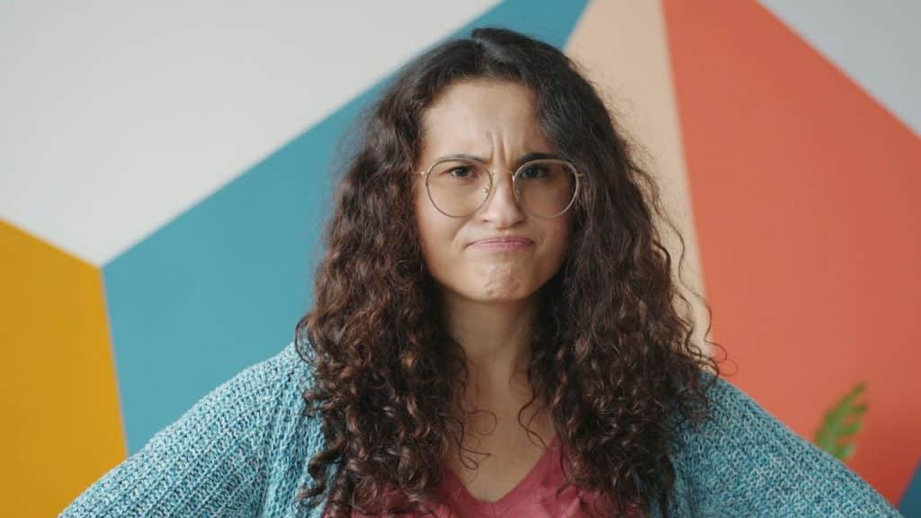 Woman with long, curly hair, glasses, and a scowling expression against a colorful geometric wall.