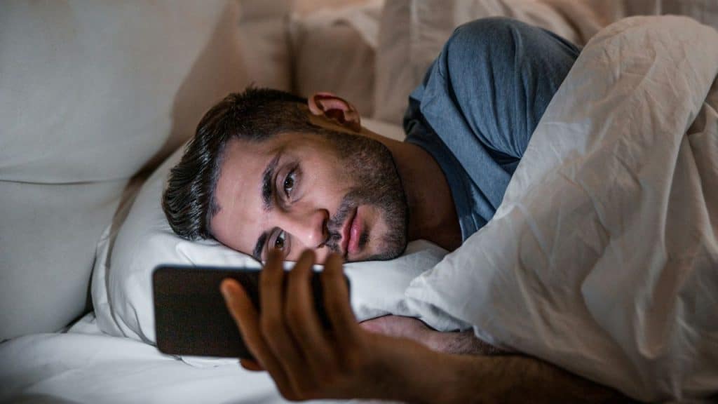 A man is lying in bed under white covers, looking at a smartphone screen he is holding.