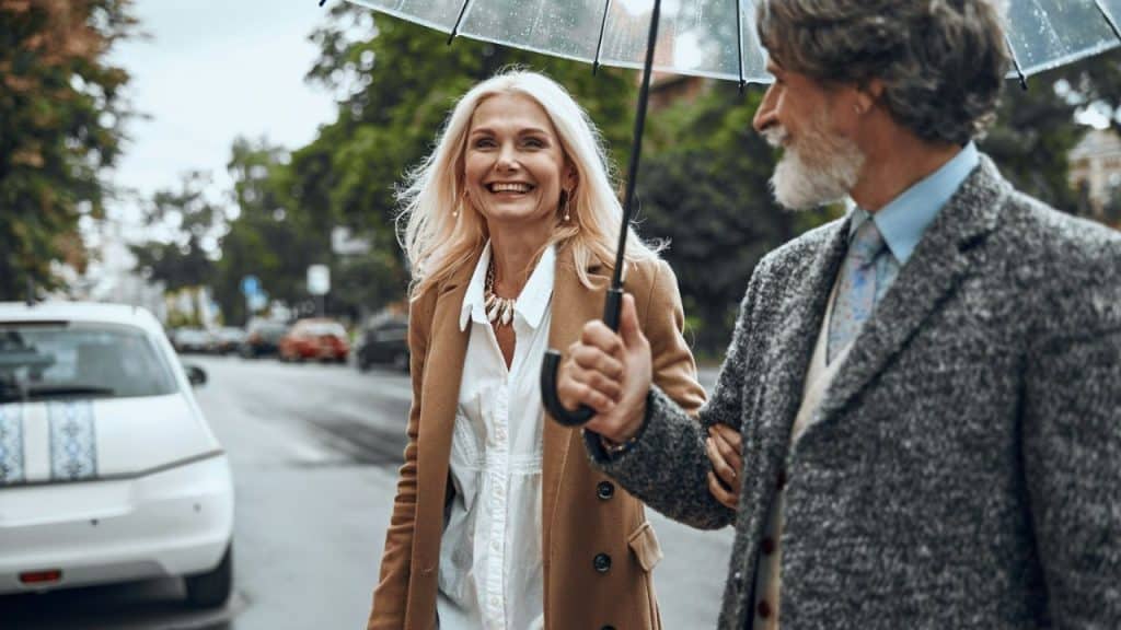 A smiling blonde woman walks arm-in-arm with a man holding an umbrella on a rainy street.