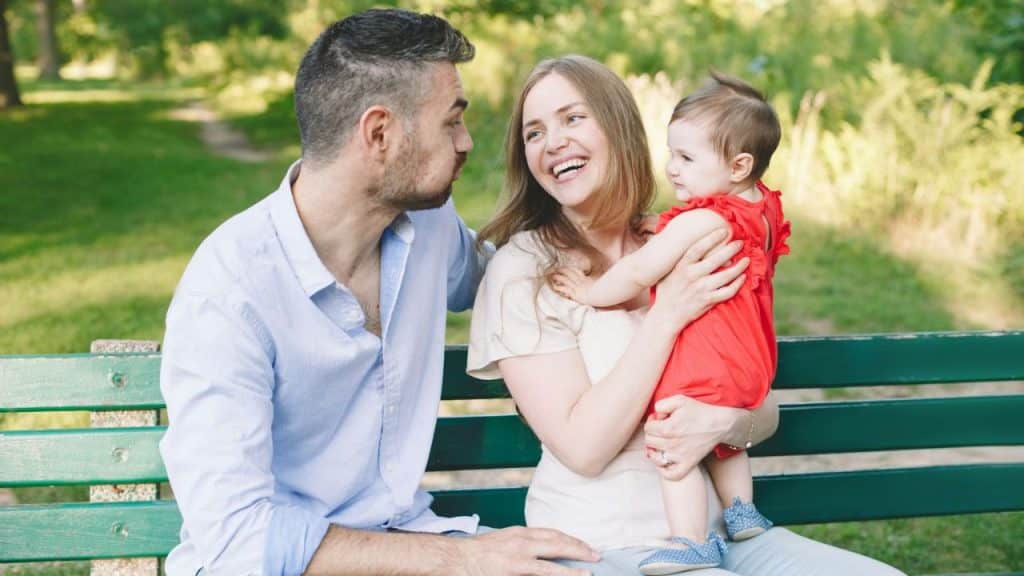 A couple on a park bench, the woman holding a smiling baby in a red outfit.