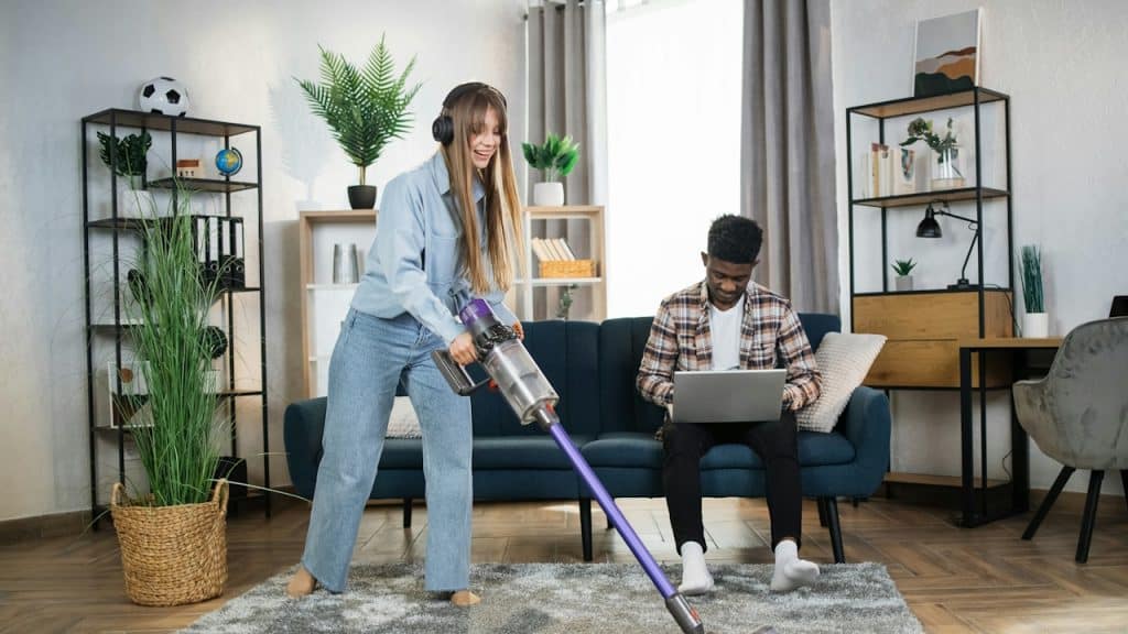 A woman is listening to music while cleaning carpet and her husband is working on a laptop.