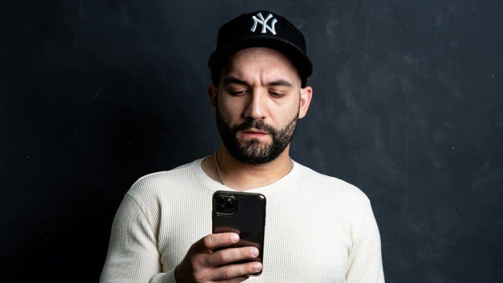 A man in a white sweater and New York Yankees hat looks down intently at his smartphone.
