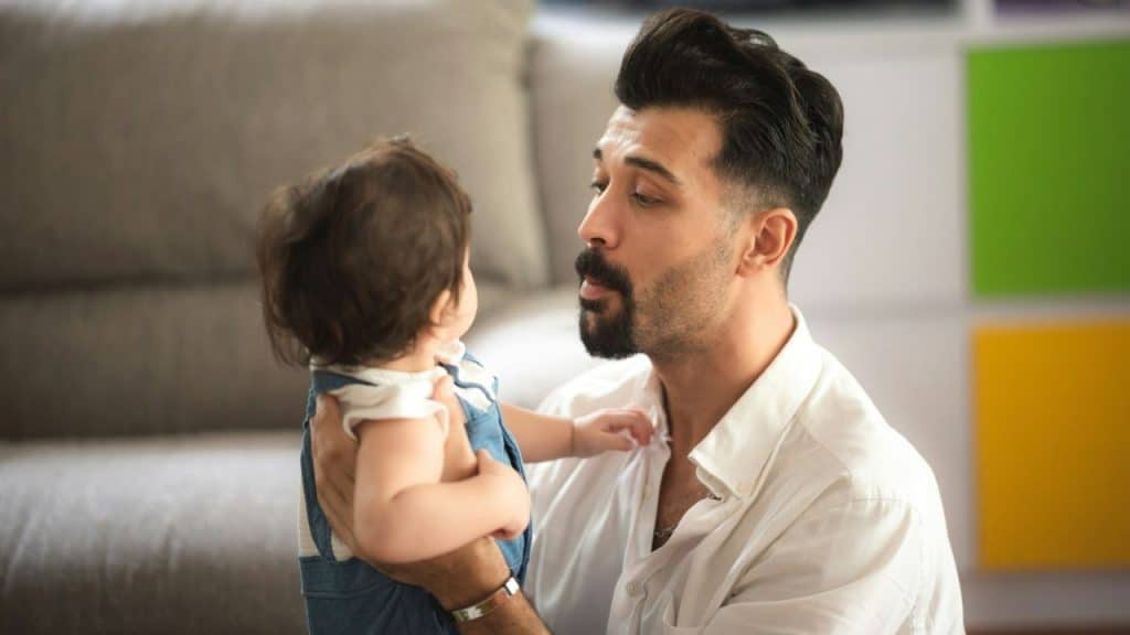 A man in a white shirt holds a baby in denim overalls and looks at the baby's face.