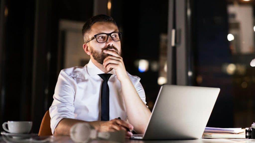 A pensive man in a shirt and tie works late at a laptop in a dark office.
