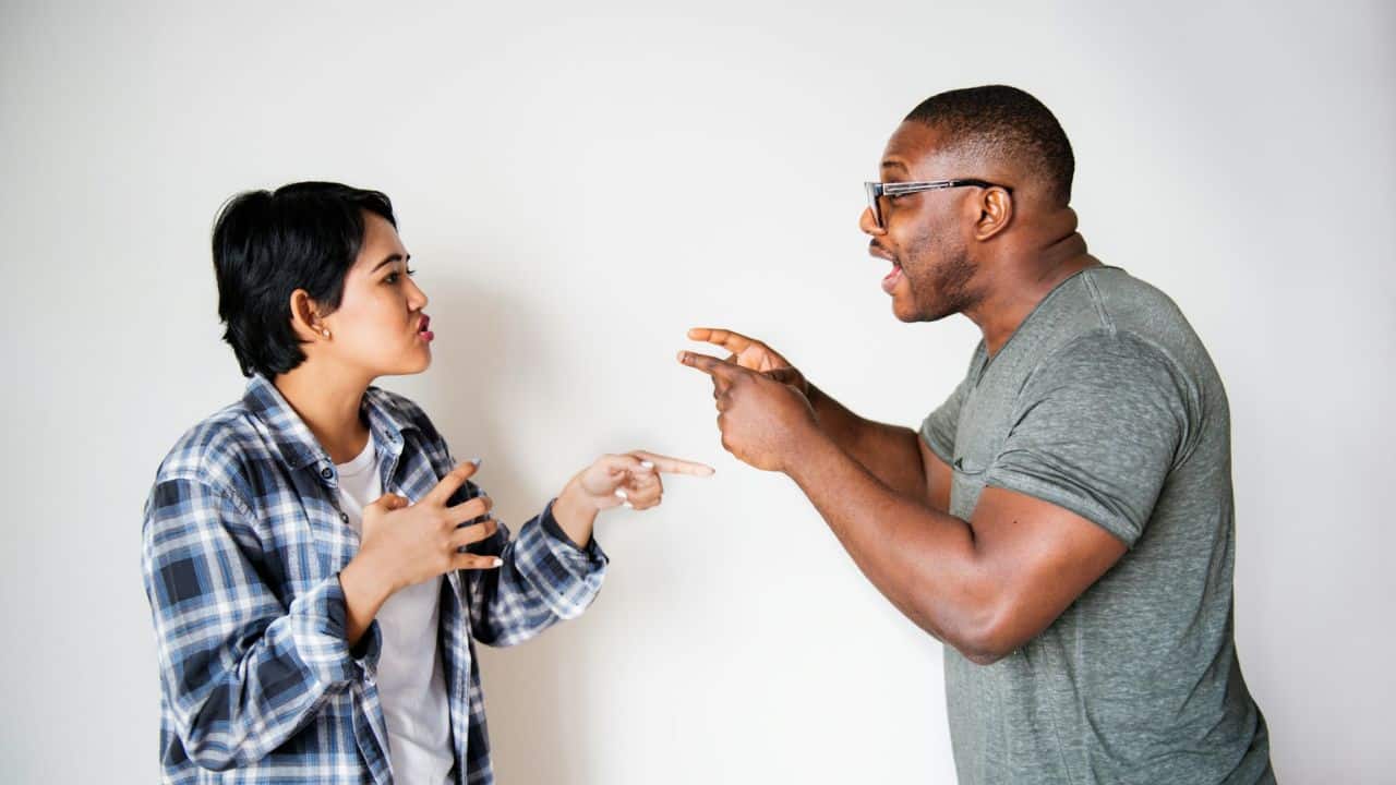 A man and woman argue, pointing fingers at each other against a white background.