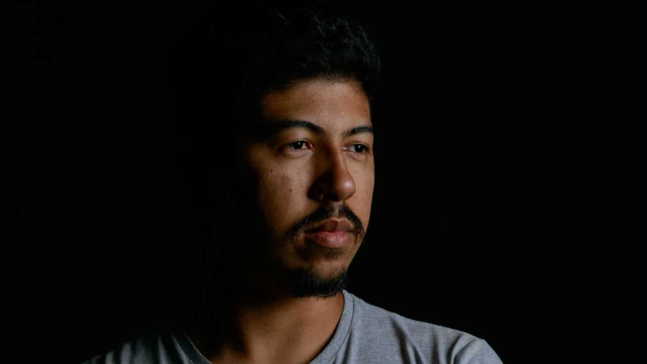 A man in a light gray shirt looks slightly to the side in a dark setting.