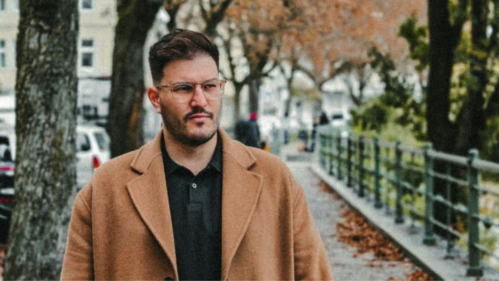 A man in glasses and a brown coat is walking on an autumnal city sidewalk.