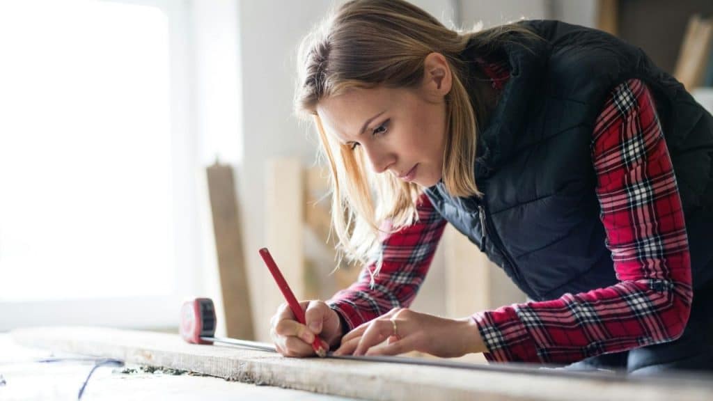 Woman wearing a plaid shirt and vest, measuring and marking wood with a pencil.