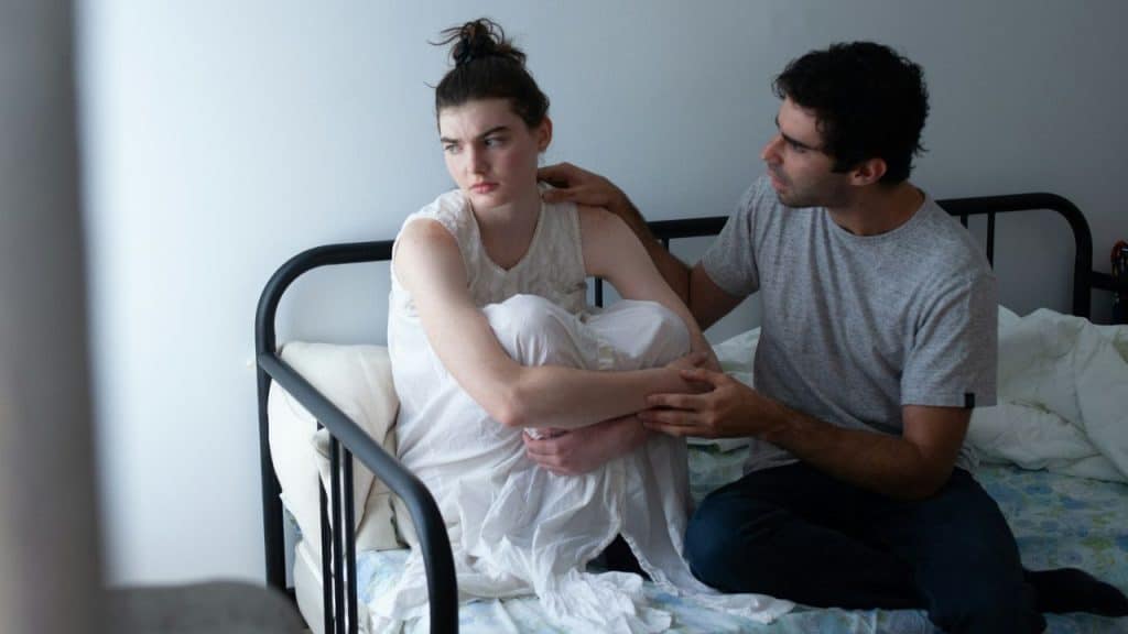 A concerned man gently touches the shoulder of a sad woman sitting on a bed.
