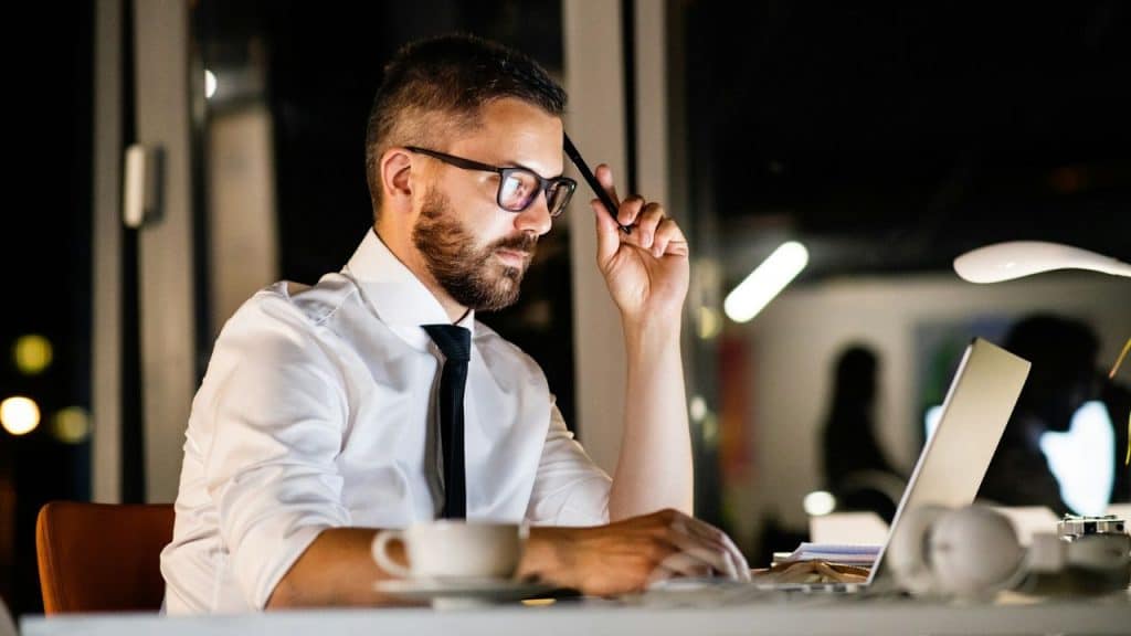 A focused man in glasses and a tie works at a laptop late at night in an office.