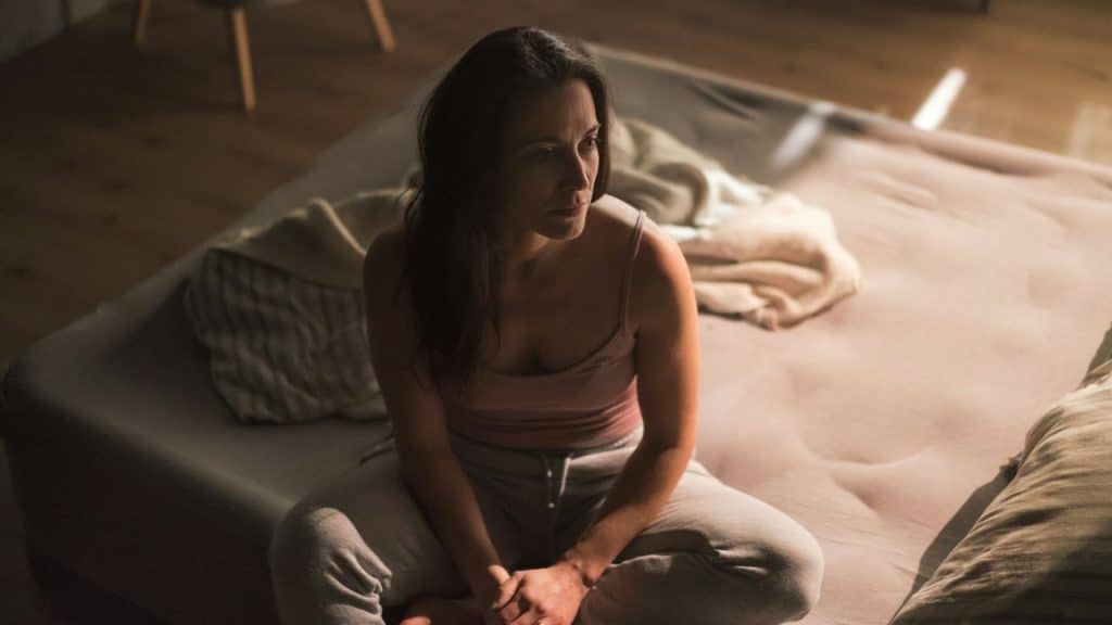 Woman with long dark hair sitting cross-legged on a mattress, looking distressed in a dimly lit room.