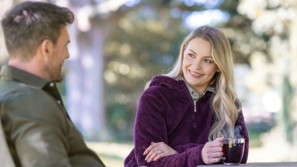 Blonde woman in a purple fuzzy jacket smiling at a man while holding a mug outdoors.