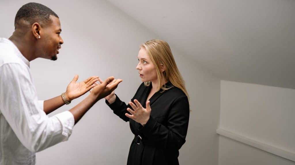 A Black man and a blonde woman are facing each other with expressive gestures.