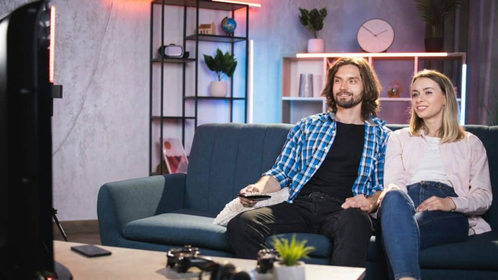 Smiling man and woman sitting on a sofa watching television in a modern living room.