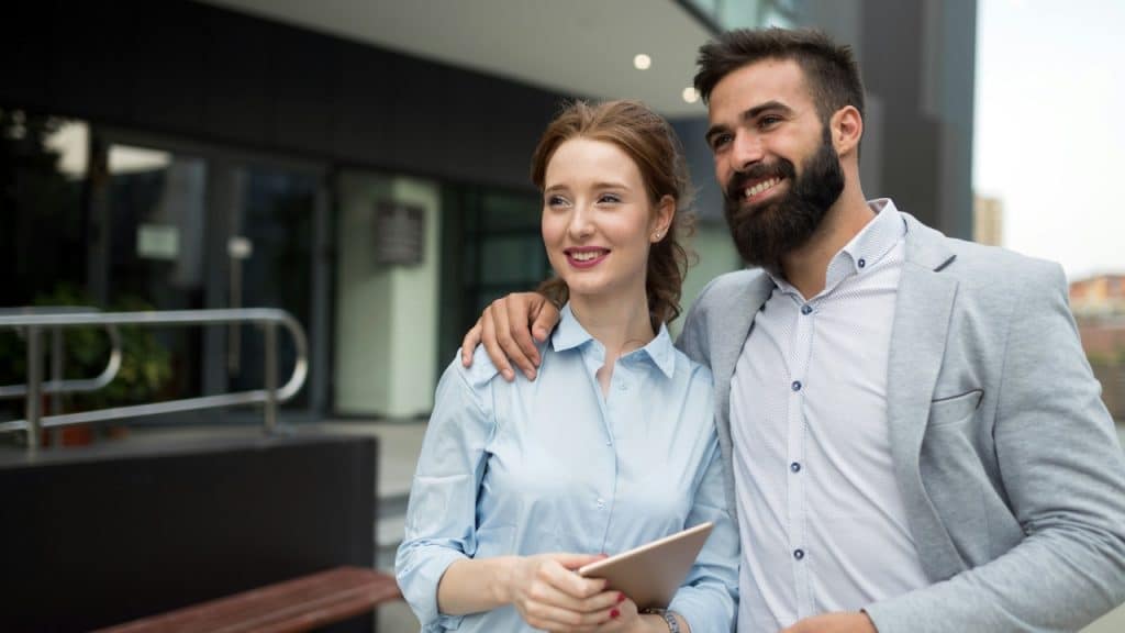 A man with a full beard and a woman with red hair are standing outside a modern building, smiling.