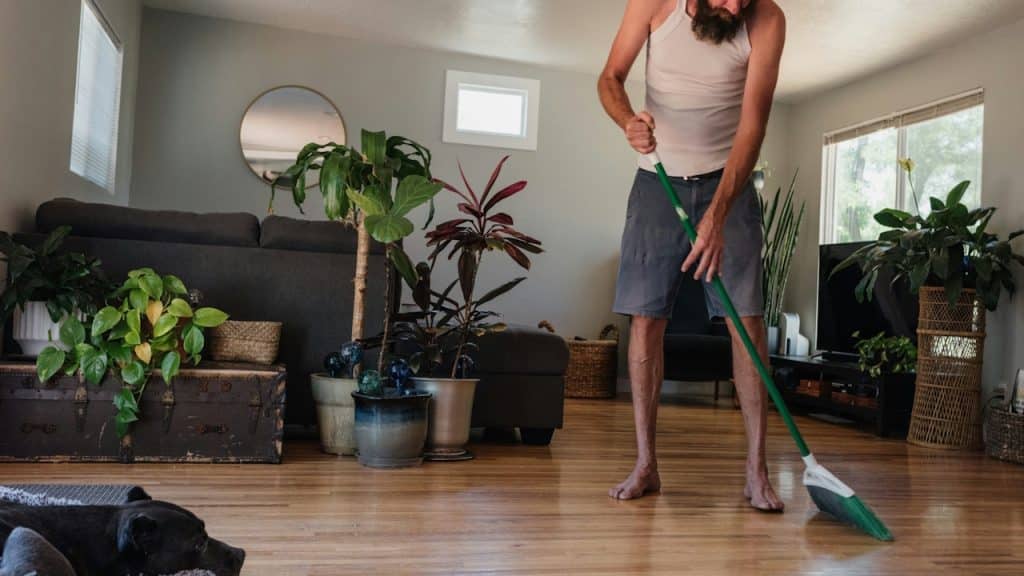 A long-haired, bearded man in a tank top and shorts is sweeping a light wood floor in a sunlit living room with many house plants.