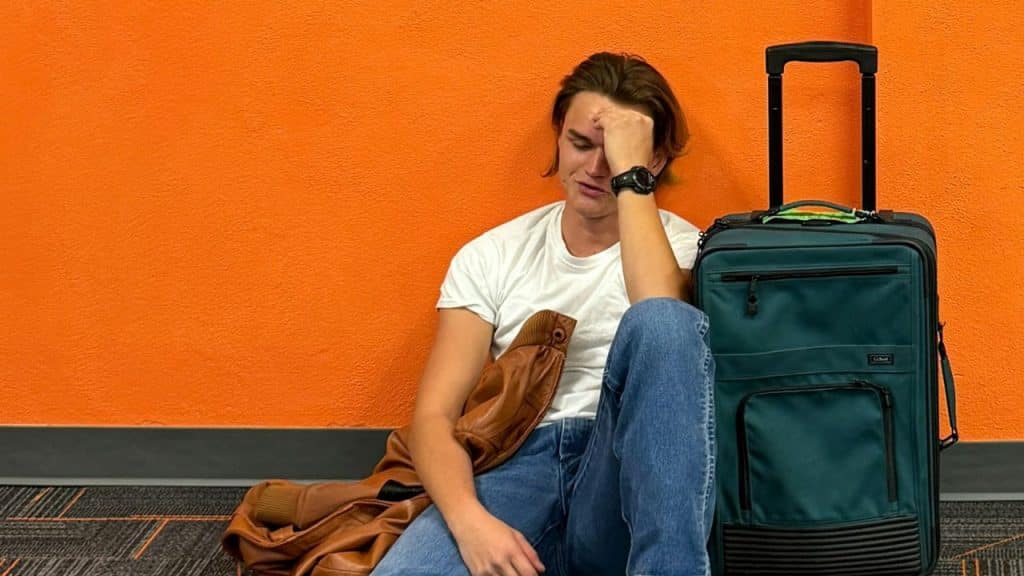 A young man is sitting on the floor, leaning against an orange wall, with a suitcase and his hand on his face.
