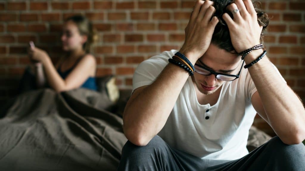 A man is sitting on a bed looking distraught, holding his head in his hands, while a woman sits in the background looking at her phone.