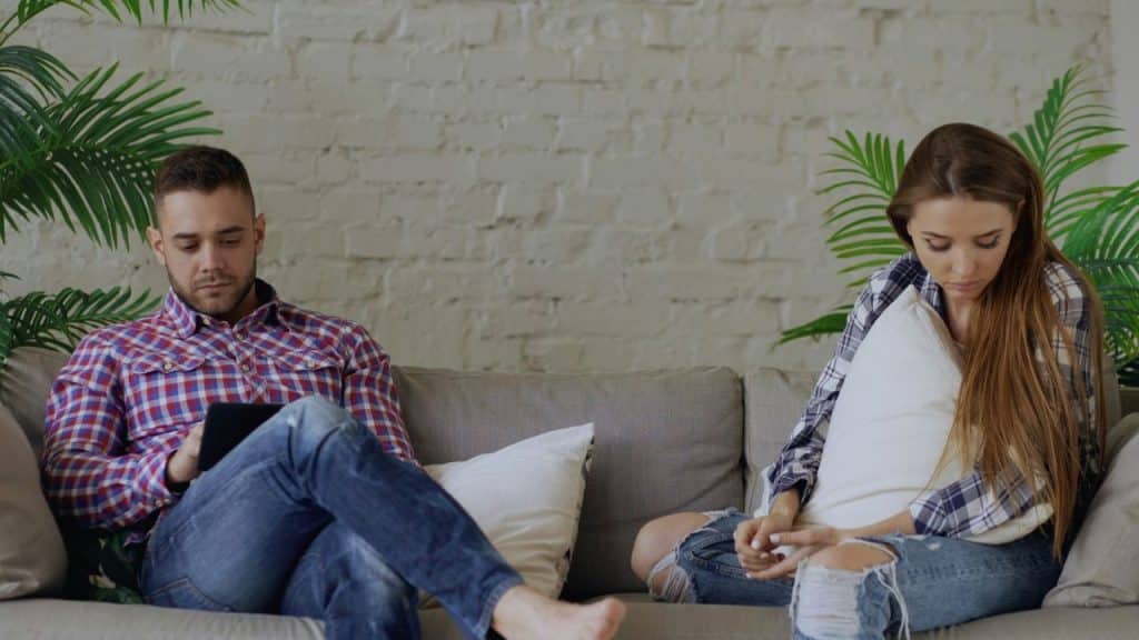 A man uses a tablet and a woman looks down, separated on a couch.