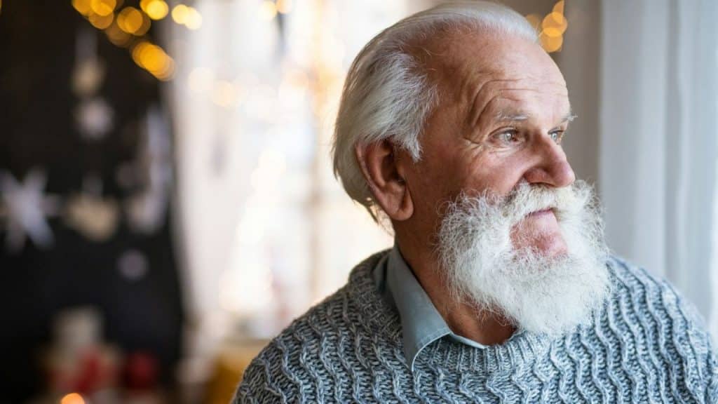 Older man with a long white beard and a gray knitted sweater looking out a window.