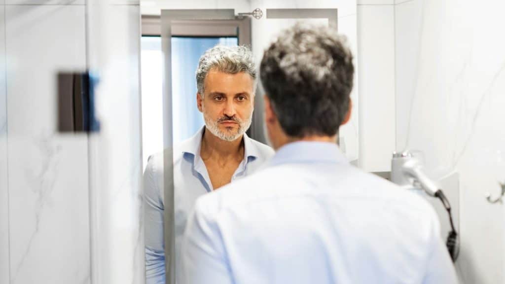 A serious, silver-haired man looks at his reflection in a mirror in a bathroom.