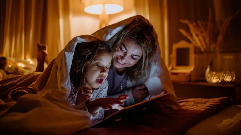 Woman and young girl under a blanket using a tablet in a dimly lit bedroom.