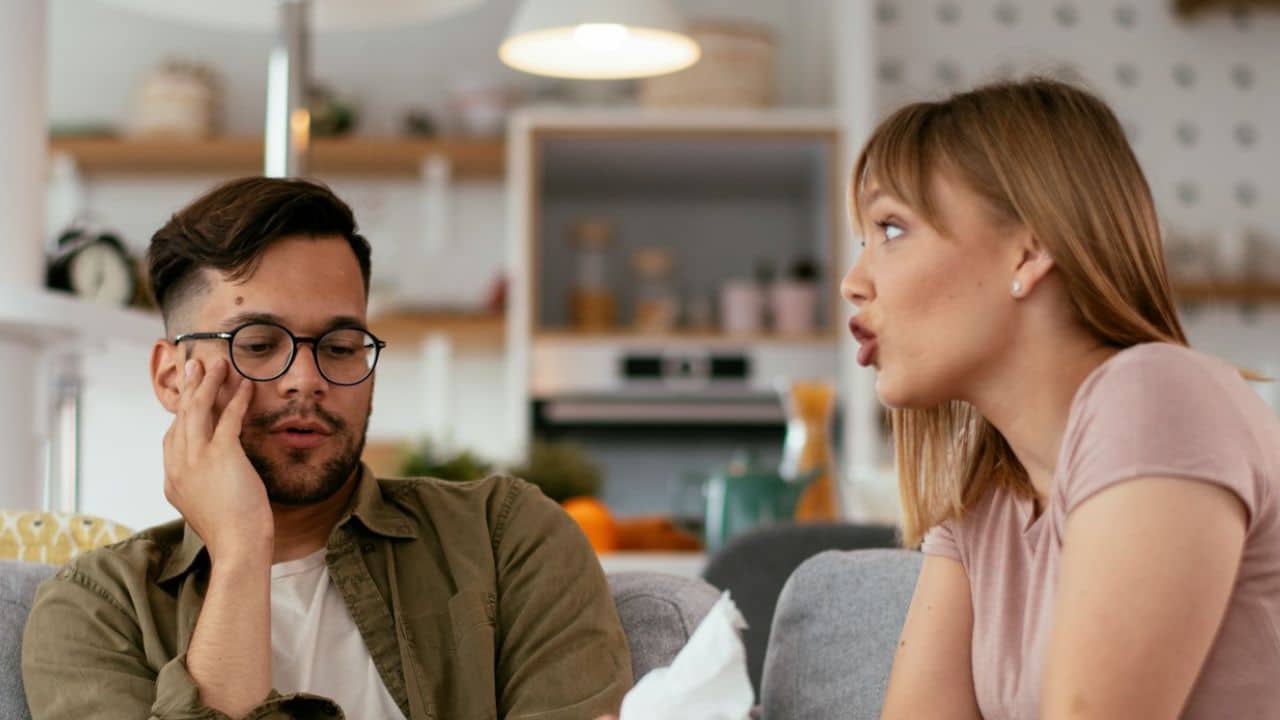 Woman speaking to a man in glasses who is resting his head on his hand on a couch.