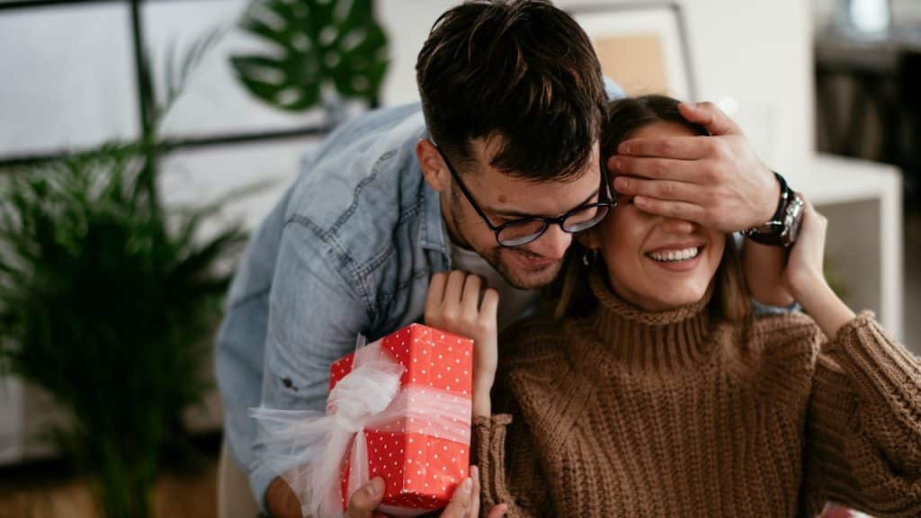 A man wearing glasses surprising a woman with a gift and covering her eyes.