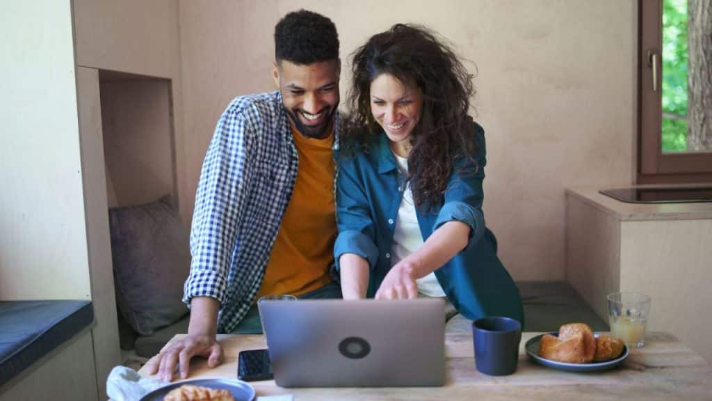A smiling man and woman look down at a laptop on a table with food and drinks.