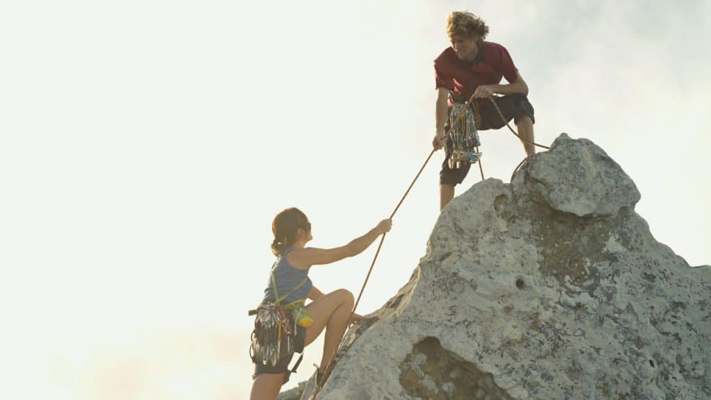 A man helping a woman climb a summit by pulling on a rope that she’s holding.