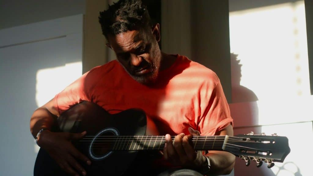 A man in an orange shirt plays an acoustic guitar indoors, illuminated by sunlight.