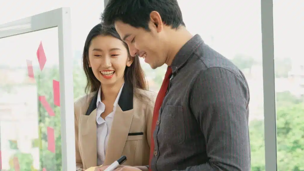 Two smiling coworkers, a man and a woman, collaborate on a whiteboard with sticky notes.