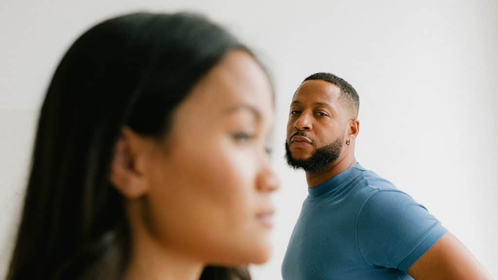 A focused shot of a serious-looking Black man in a blue shirt looking directly at the camera while a woman's blurred profile is in the foreground.