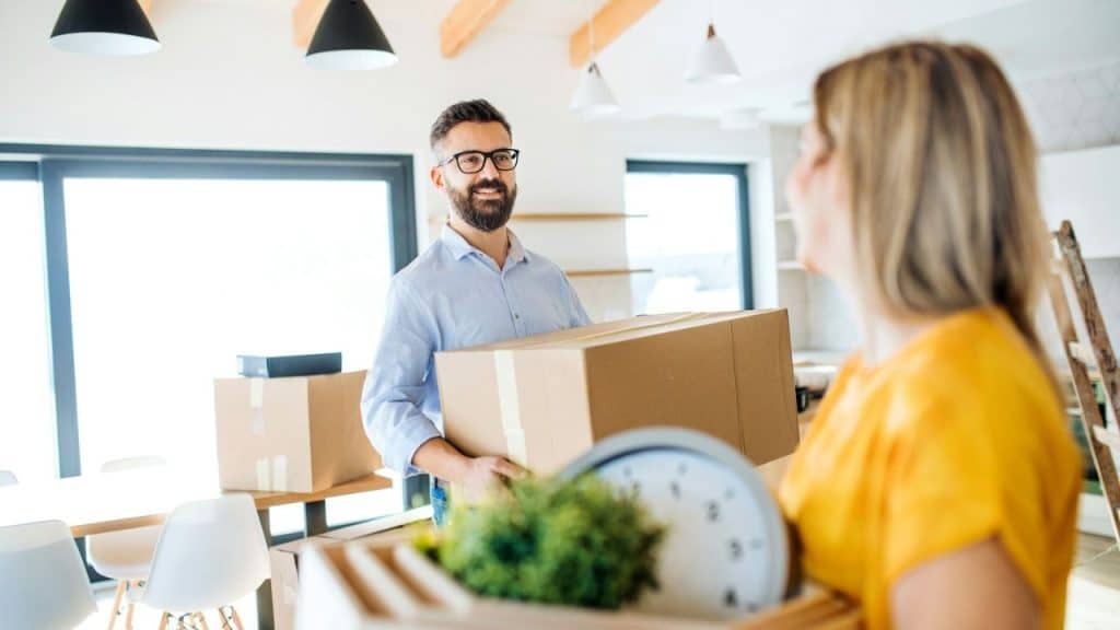A smiling, bearded man carries a cardboard box and looks at a woman carrying a box in a bright, modern room.