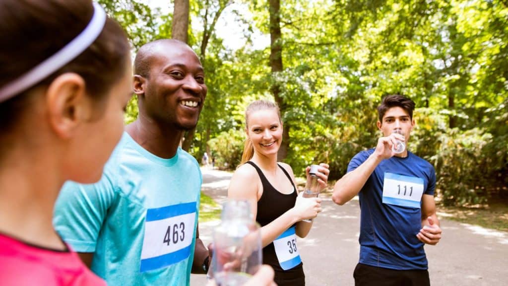 Four happy people wearing race bibs and athletic clothing are taking a water break outdoors after a run.