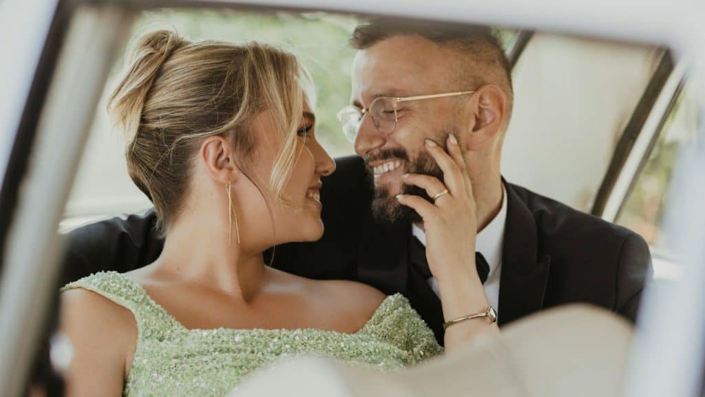 A couple smiles and looks at each other while sitting in a car's back seat.